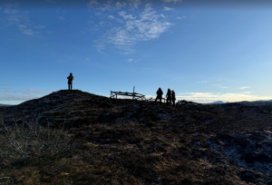 4 personer står på en haug og ser på nedbrente forskningsstrukturer. Det er spor av snø på bakken og blå himmel.